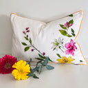 A rectangular ivory white decorative throw pillow with pink, white, and yellow flowers and green leaves, finished with a thin orange-yellow piping border. The pillow is styled on a white surface with yellow and pink flowers 