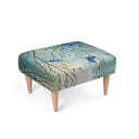 A rectangular upholstered footstool with a printed design of  splendid  fairy wren blue birds perched on tree branches with teal and beige leaves, supported by four light wood legs on a white background.