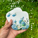 A hand holds two square coasters with rounded corners featuring a blue and white floral design, set against a blurred green grass background.
