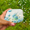 A hand with pink nail polish holds a 2 square wooden coasters with a glossy finish decorated with a blue and white floral print, set against a blurred background of green grass and small white flowers.