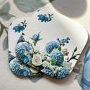Two square coasters with rounded corners featuring a floral print of blue hydrangeas, white roses, and green leaves on a white background, styled on a textured light surface with soft shadows.