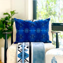A cobalt blue throw pillow with a light blue geometric snowflake pattern, styled on a beige armchair in a bright, modern room with a green plant in the background.