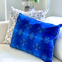 A vibrant cobalt blue rectangular throw pillow with white snowflake patterns rests on a cream-colored sofa next to an ivory and black patterned accent pillow in the background.
