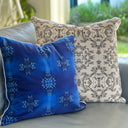 Two decorative throw pillows on a blue sofa in a real indoor setting with blurred greenery in the background. The front pillow is cobalt blue velvet with a lighter blue snowflake-like pattern and white piping.