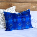 A cobalt blue rectangular throw pillow with a grey snowflake pattern, styled on a bed with white sheets and other decorative pillows in front of a wooden headboard.