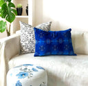 A vibrant cobalt blue throw pillow with white snowflake patterns rests on a cream-colored sofa next to a white and blue floral ottoman, with a patterned accent pillow in the background.