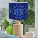 A table lamp with a cylindrical natural wood base and a matching drum shade in cobalt blue, decorated with a light blue snowflake pattern. The lamp is styled on a tabletop with green houseplants and neutral decorative blocks in the background.