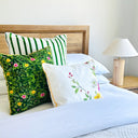 Three decorative throw pillows arranged on a bed with white bedding, a wooden headboard, and a bedside lamp. The pillows include a green and white striped velvet  pillow, the Forest green floral velvet pillow with yellow and pink flowers, and a white Lily Cotton linen floral pillow with green leaves and pink lilies. 