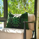 An emerald green velvet cushion with snowflake shaped  floral pattern, resting on a beige armchair in front of a window with outdoor green foliage visible.