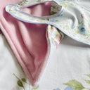 A close-up of a baby blanket. with a soft pink plush back and a white border printed with a delicate pastel floral pattern, folded back on the front of white floral blanket