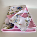 A baby gift set featuring a white bib with a blue and green floral print and a pink fleece lining, displayed on top of folded matching pink and floral blankets on a white background.