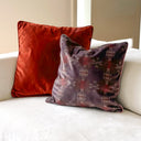 Two velvet throw pillows on a white sofa. The back pillow is a solid rust red, and the front pillow is a dark brown with a repeating light beige floral pattern.