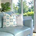 A light blue leather sofa in a bright room, styled with two decorative throw pillows: one white with a blue ornamental pattern and one solid white.