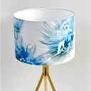 A table lamp with a white drum shade featuring a blue blushing bride floral motif, supported by a gold tripod base against a light gray background.