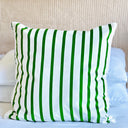 A square throw pillow in velvet with vertical green and white stripes, resting on a bed with white pillows and a beige textured headboard in the background.