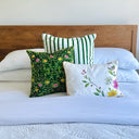 Three decorative throw pillows styled on a bed with white bedding and a wooden headboard. The pillows include a green and white striped pattern, a dark green floral print with yellow and pink flowers, and a white pillow with a pink and yellow floral design.