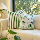 Two decorative throw pillows on a cream sofa in a bright living room. One pillow has bold green vertical stripes on a white background, and the other has a white with green eucalyptus leaves. 