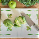 A stonemax cutting board with a green leaf pattern, shown on a wooden table with Brussels sprouts, a green apple, and a chef's knife.