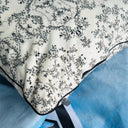A close-up of a decorative pillow with an off-white background and a delicate black floral vine pattern, finished with black piping and a small silver metal zipper pull.