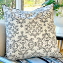 A square decorative throw pillow with a black damask-style floral pattern on an off-white background, featuring black piped edges, resting on a light-colored armchair near a window with a plant in the background.