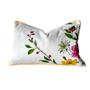 A rectangular ivory white throw pillow with a vibrant floral print featuring pink, white, and yellow lilies with green leaves, finished with a thin yellow piping border.