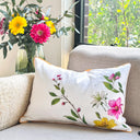 A rectangular ivory white throw pillow with a colorful floral print of pink, yellow, and white flowers with green leaves, resting on a beige textured sofa next to a vase of fresh flowers.
