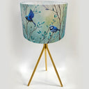A table lamp with a cylindrical drum shade printed with blue birds and leafy branches, supported by a gold-finished tripod base on a white background.