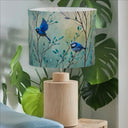 A table lamp with a cylindrical light wood base and a drum shade printed with two blue birds perched on branches against a soft blue-green background, styled beside green houseplants next to a light blue sofa.