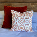 Two decorative throw pillows on a bed: a front square pillow with terracotta and white damask pattern on a white background, and a rear rectangular rust-red velvet pillow against a wood headboard.