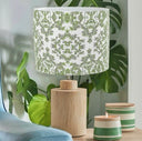 A table lamp with a white drum shade featuring a green damask pattern and a natural wood cylindrical base, styled on a wooden table next to two green and white striped candles and a blue chair.