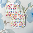 Two square white coasters with rounded corners, decorated with a colorful floral pattern of pink, blue, orange, and yellow flowers, placed on a light blue floral surface.