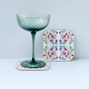 A green ribbed stemmed glass and a square white floral coaster with colorful floral design, arranged on a light gray studio background.