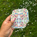 A hand holds 2 square round edged  coasters with a colorful floral pattern against a blurred green grassy background with small white flowers.