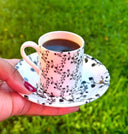 A hand with red nail polish holds a white bone cup and saucer decorated with a black floral branch pattern, filled with coffee, against a blurred green grass background.